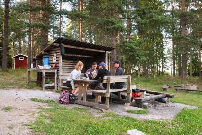 Four campers sit at picnic table in front of campfire shelter in Pitea, Sweden